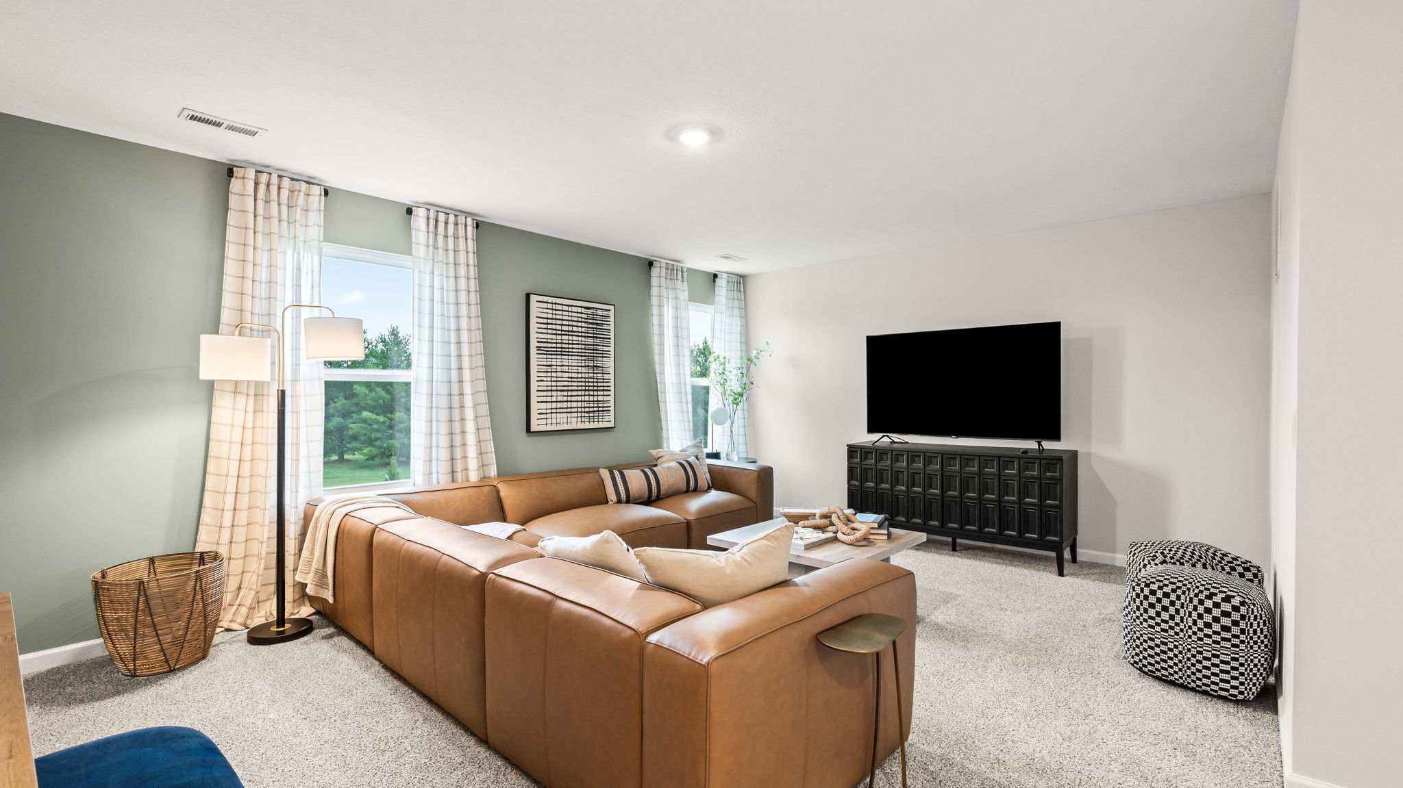 Upstairs living room with a tan sectional sofa, green accent wall, tall windows with checked curtains, floor lamp, coffee table, television on a dark media console, and patterned accent ottomans.