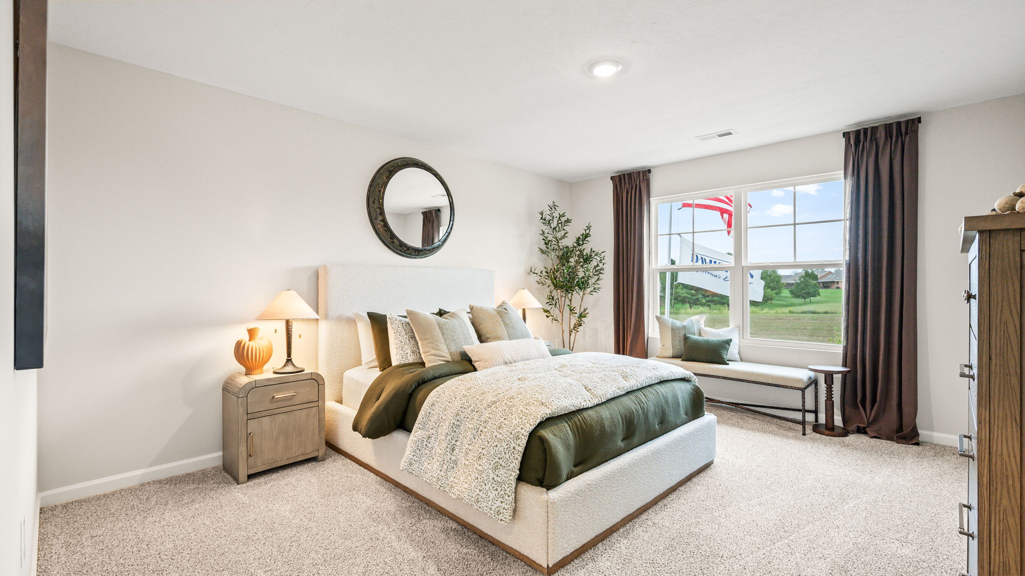 Bedroom with a white upholstered bed, green and cream bedding, matching nightstands and lamps, round wall mirror, tall curtained window, bench, decorative tree, and carpeted flooring.