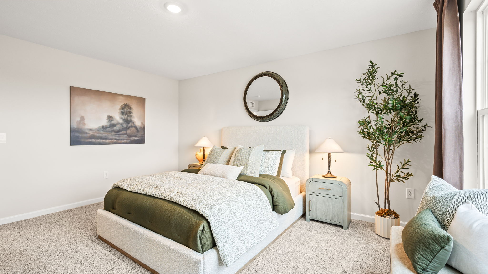 Bedroom with a white upholstered bed, green bedding, matching bedside lamps, round wall mirror, landscape artwork, decorative tree, and a chair near the window.