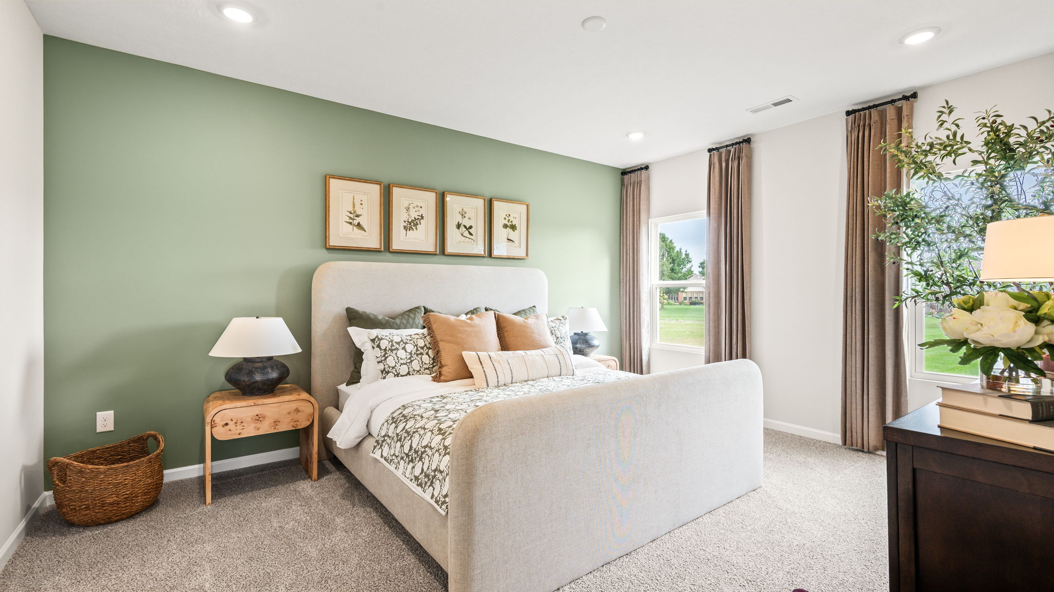 Bedroom with an upholstered bed against a green accent wall, green and neutral bedding, four botanical prints, bedside lamps, tall curtained windows, and a dark dresser.