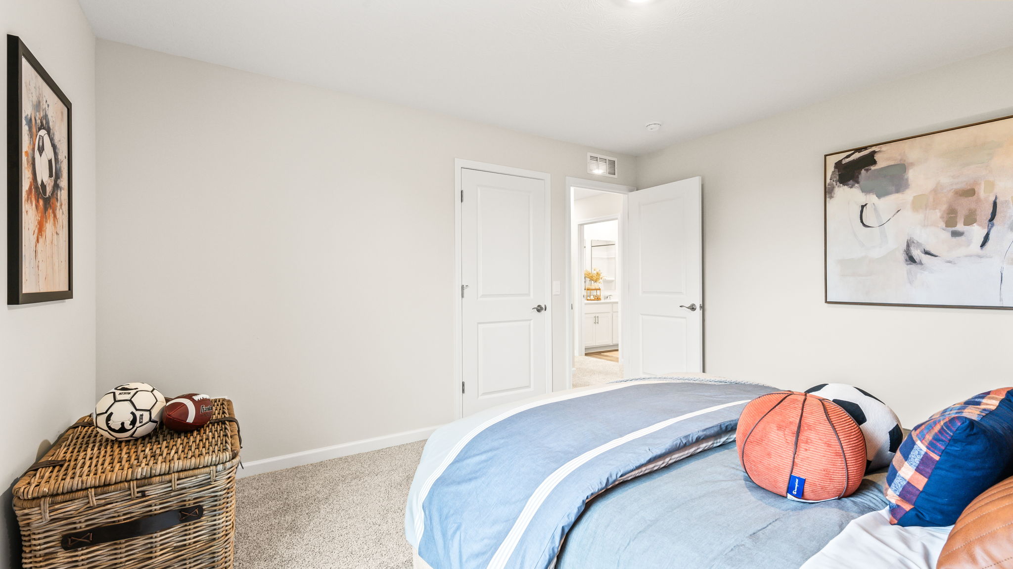 Bedroom viewed from beside the bed, with sports-themed pillows, a woven storage chest holding balls, neutral walls, framed artwork, and open doors to the hallway and bathroom.