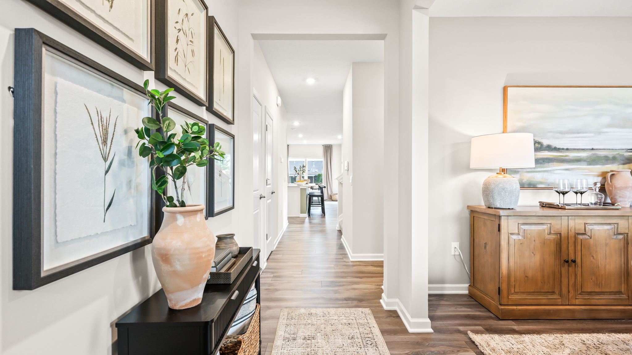Entry hallway with a black console table, framed botanical artwork, ceramic vase, wood-look flooring, runner rug, and an opening to a dining room with a wood sideboard.
