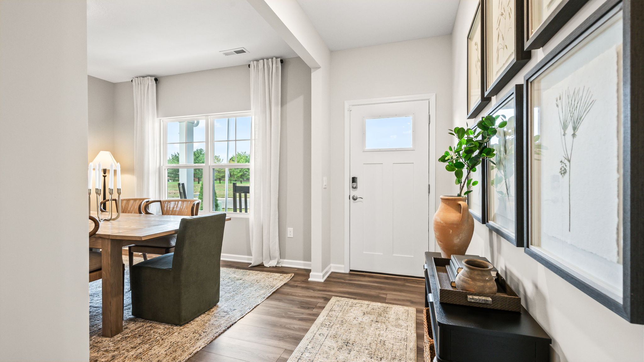 Front entry with a white door, wood-look flooring, runner rug, black console table with framed art and a vase, and an adjacent dining room with a large window.