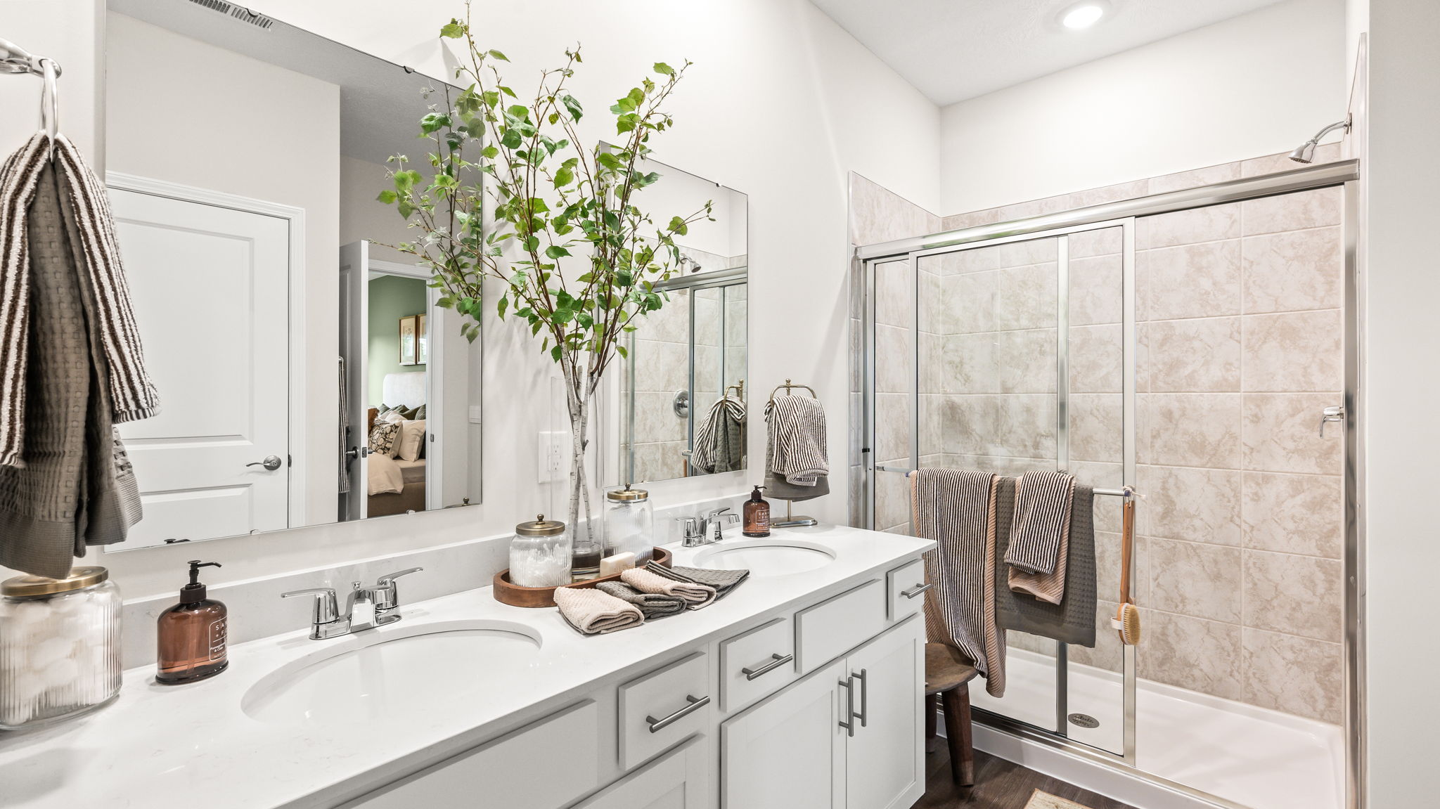Bathroom with a long white double-sink vanity, large mirrors, chrome faucets, decorative branches, striped towels, and a glass-enclosed beige tile shower.
