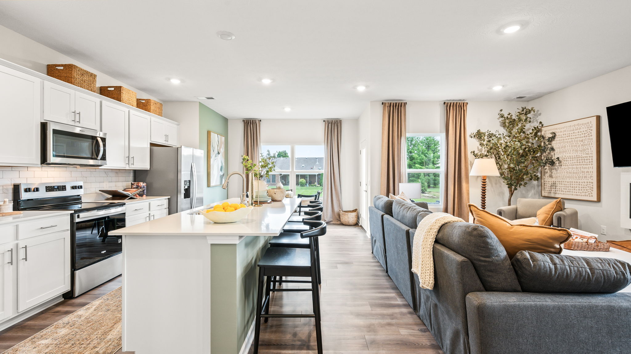 Wide open-plan kitchen and living area with white cabinetry, stainless steel appliances, a long pale green island with black stools, gray sectional seating, tall curtained windows, and a fireplace.