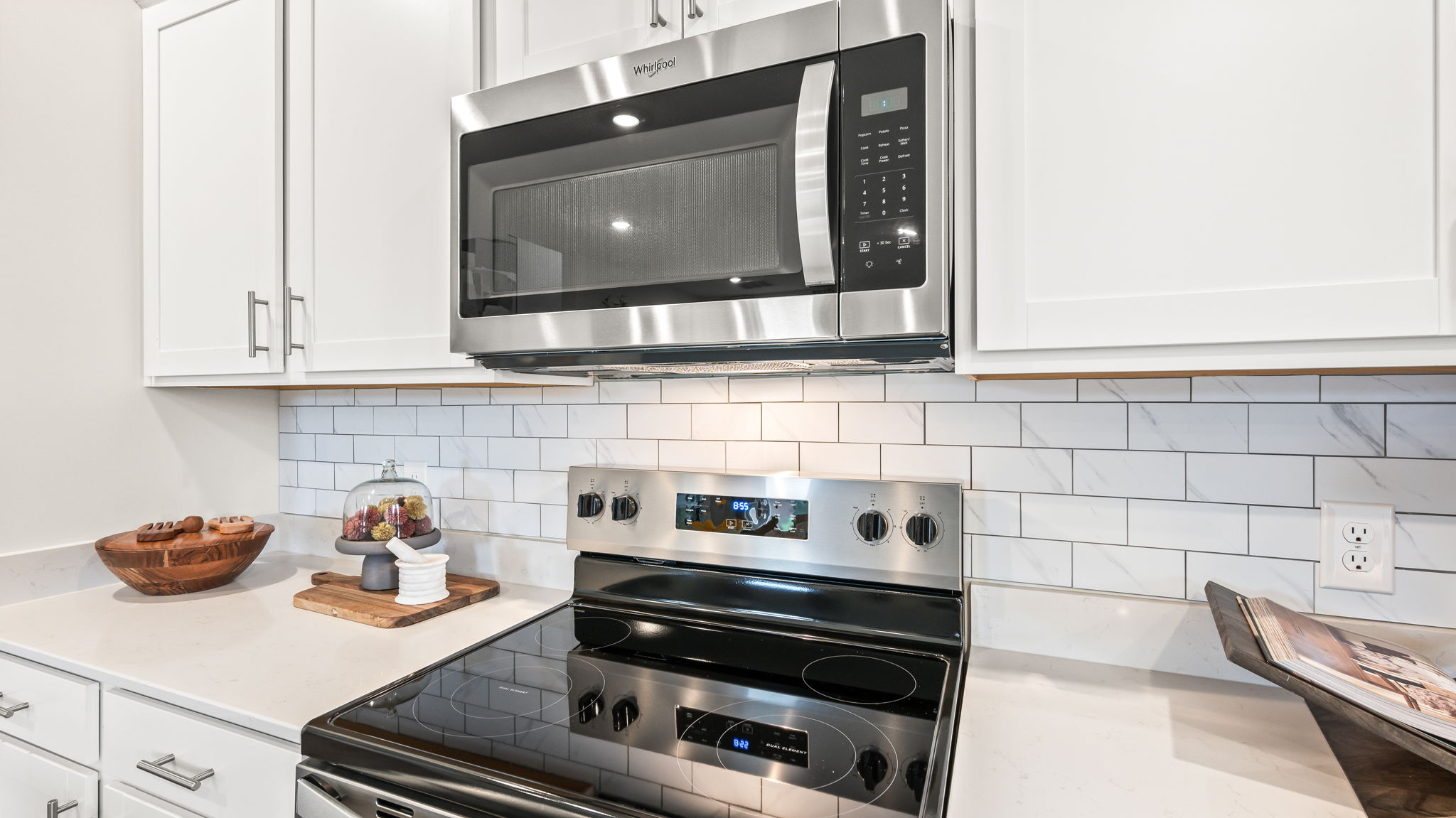 Close view of a stainless steel microwave above an electric range, surrounded by white shaker cabinets, white countertops, and a white marble-look subway tile backsplash.