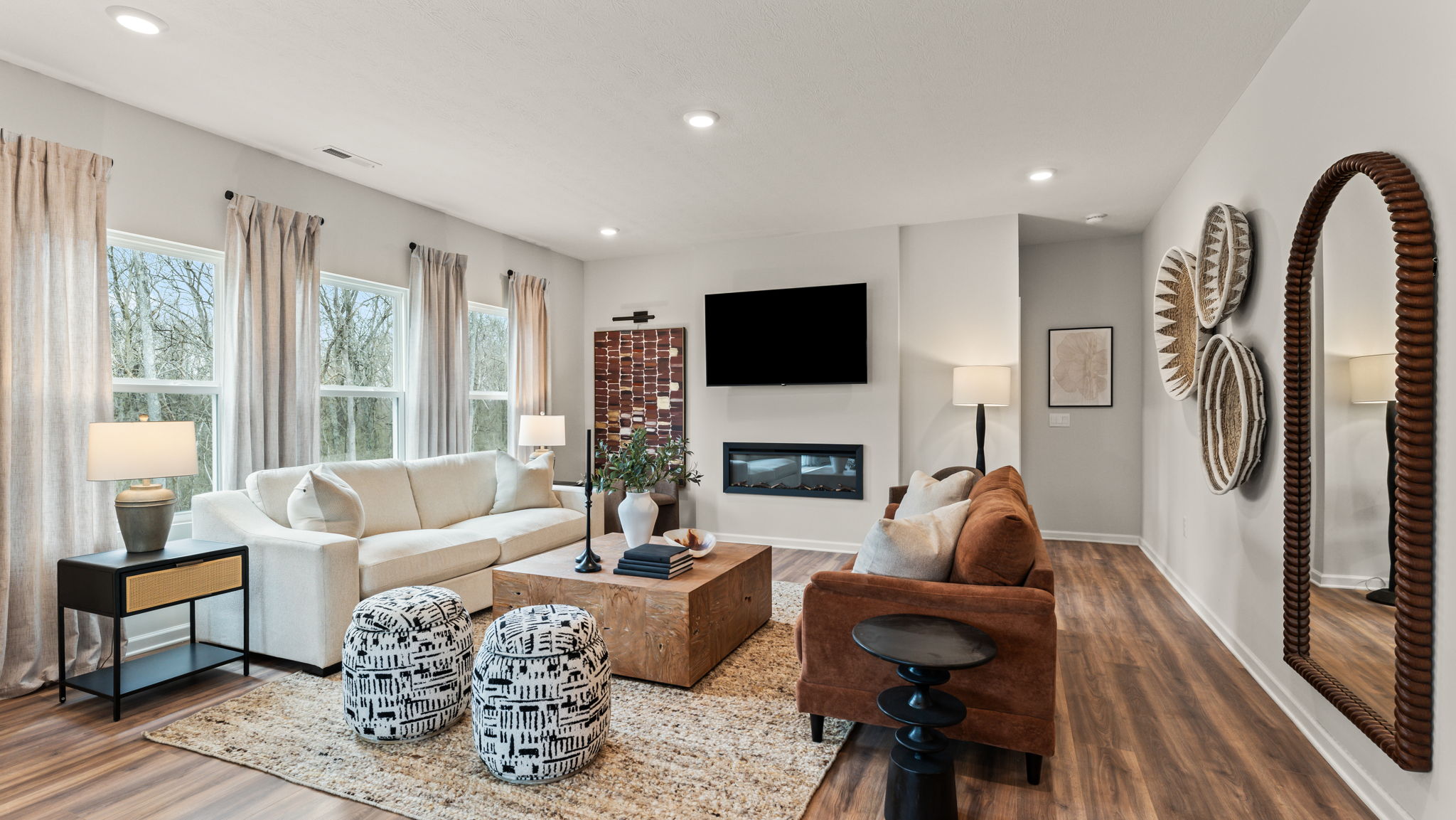 Modern spacious great room with a mix of warm ambient lighting and natural light from large windows adjacent to long drapes. Brown furniture and art displays on the wall are broken up by white accents