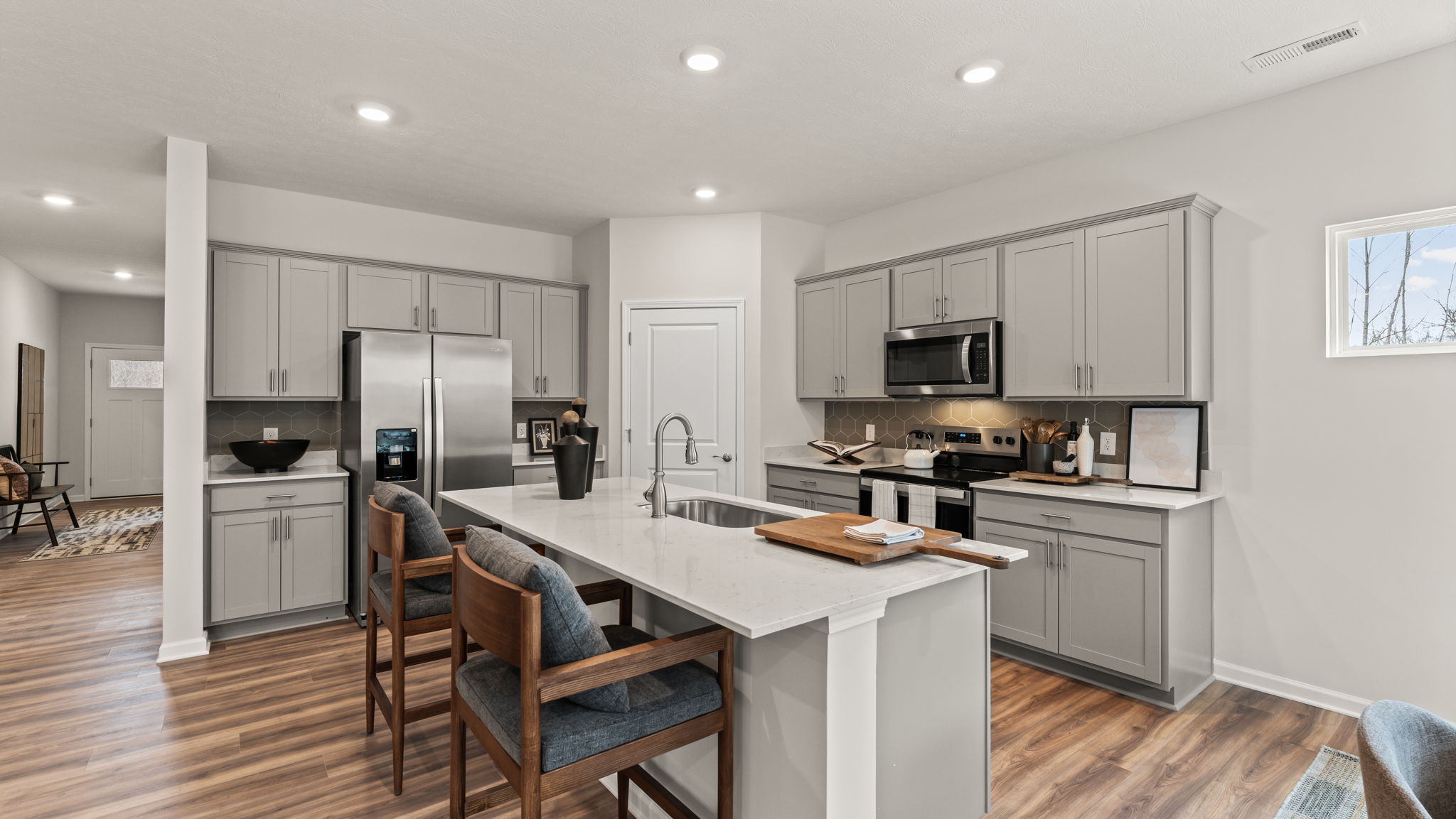 Wide angle three-quarter perspective image of a modern open-concept kitchen with gray cabinets, stainless steel appliances, island, and brown wood-accented chairs