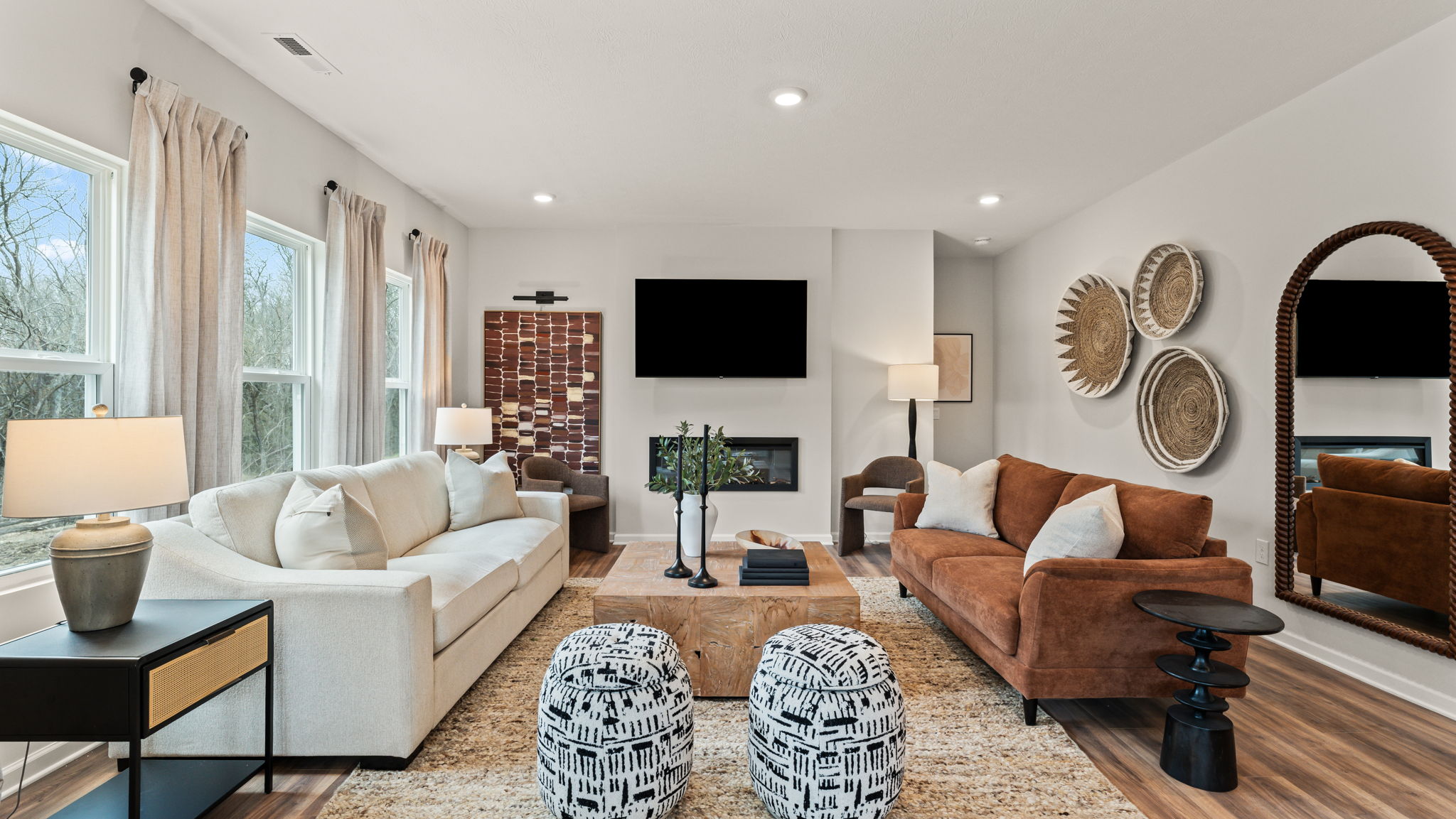 Modern spacious great room with a mix of warm ambient lighting and natural light from large windows adjacent to long drapes. Brown furniture and art displays on the wall are broken up by white accents
