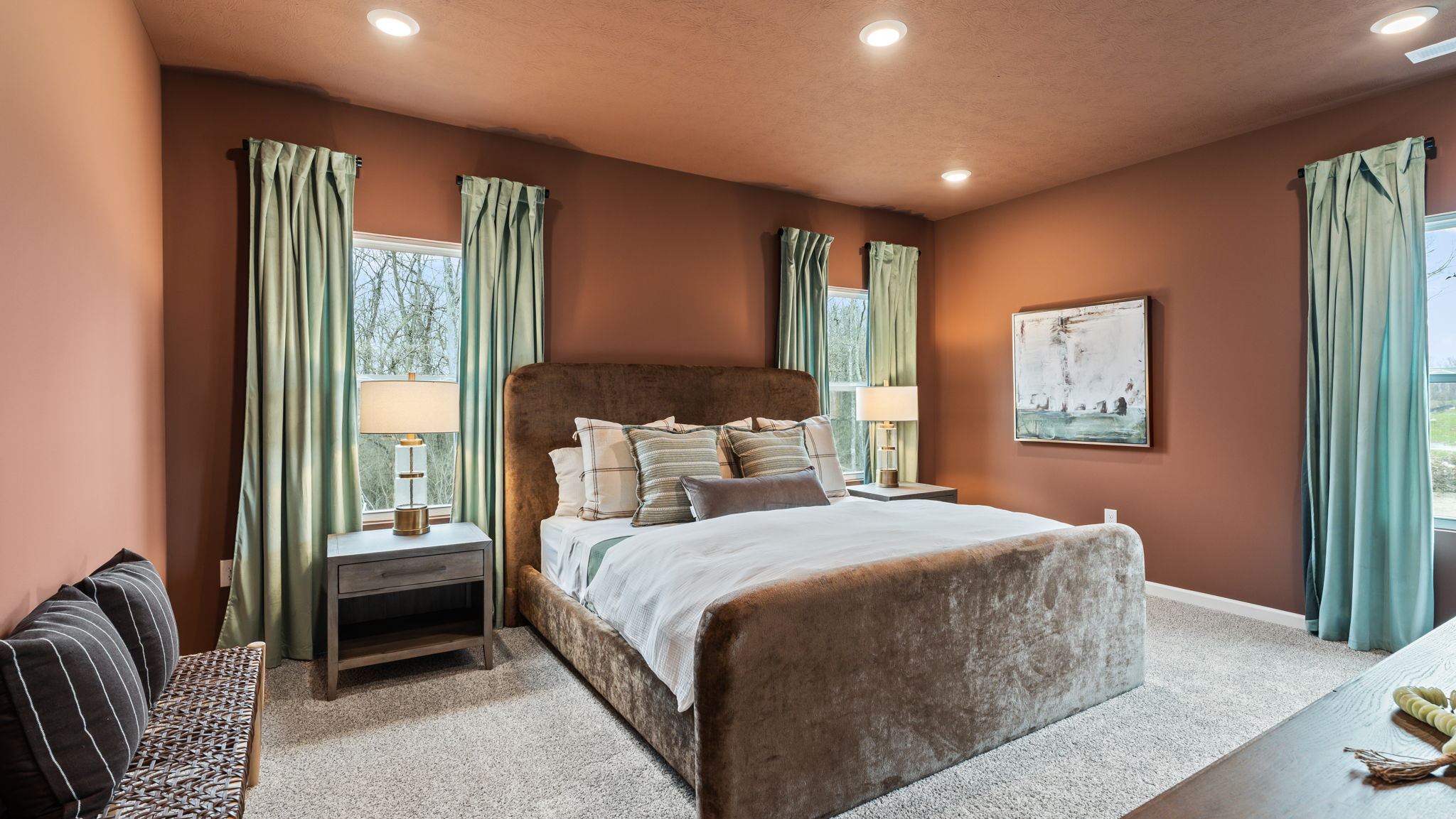 Large primary bedroom suite with suede bed frame, warm ginger walls contrasted by lush green drapery, and a braided bench with wooden legs.