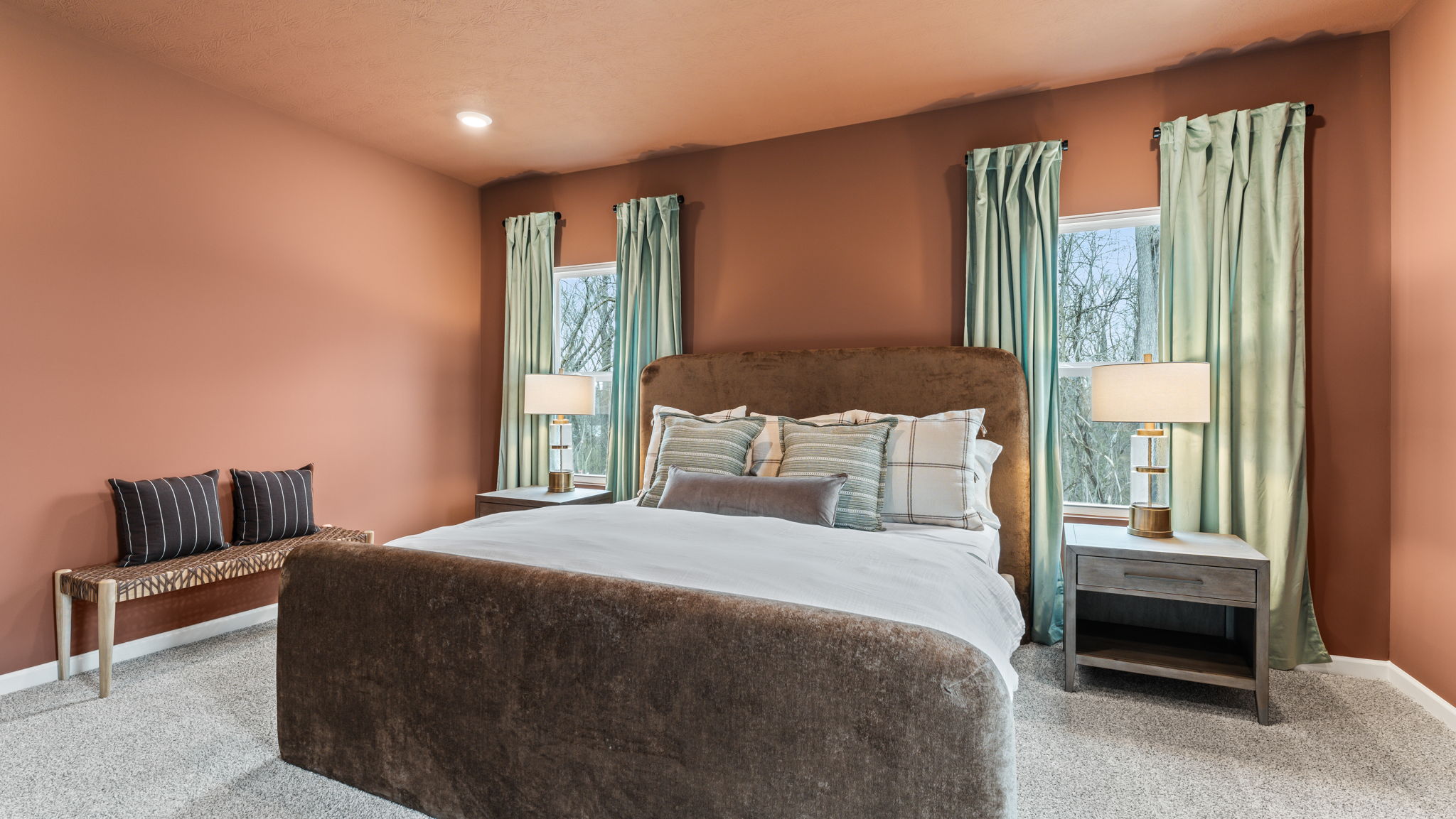 Large primary bedroom suite with suede bed frame, warm ginger walls contrasted by lush green drapery, and a braided bench with wooden legs.