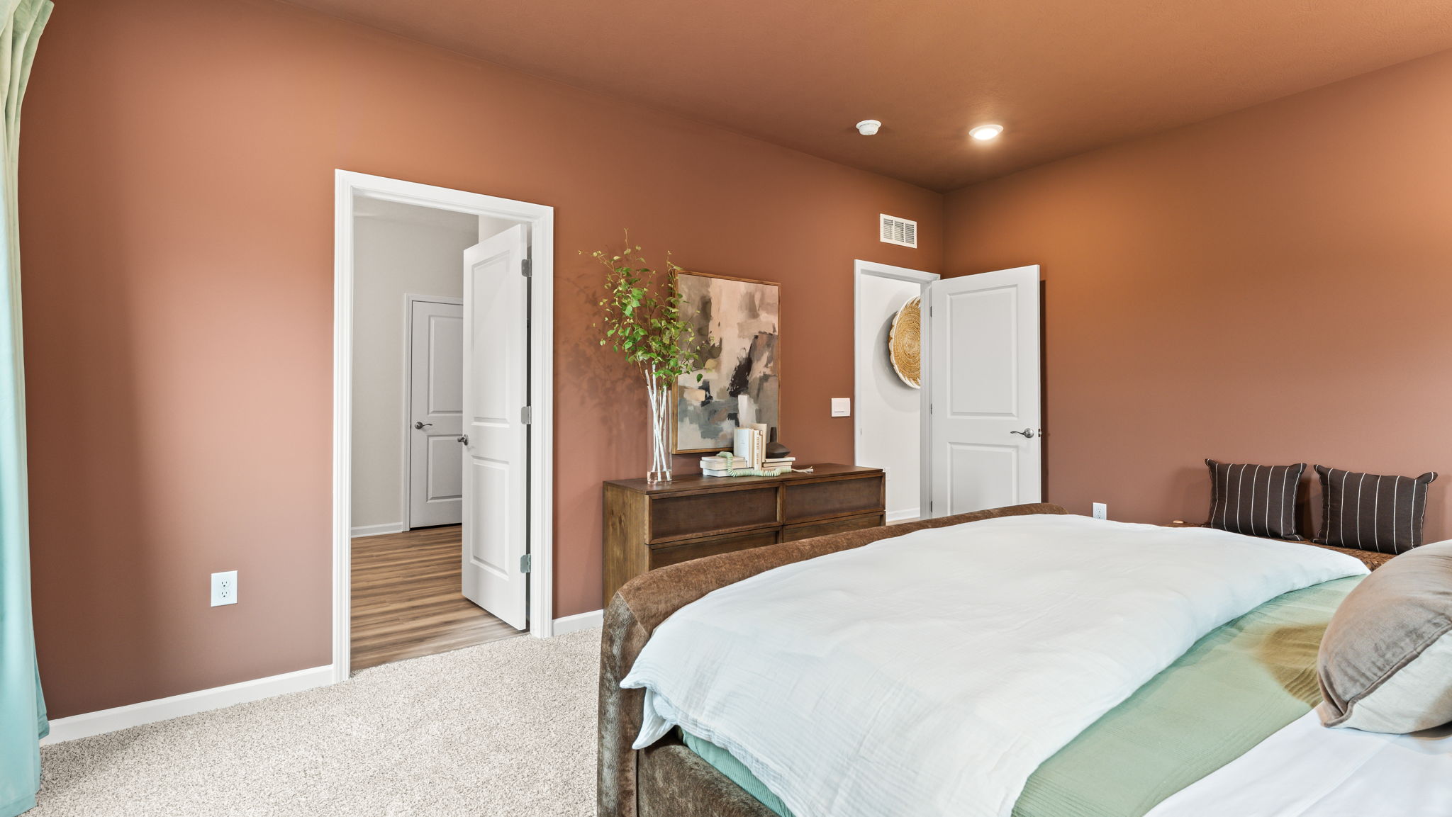 wide image of inviting primary bedroom suite from the bedside, with dresser-drawer and door to primary bathroom