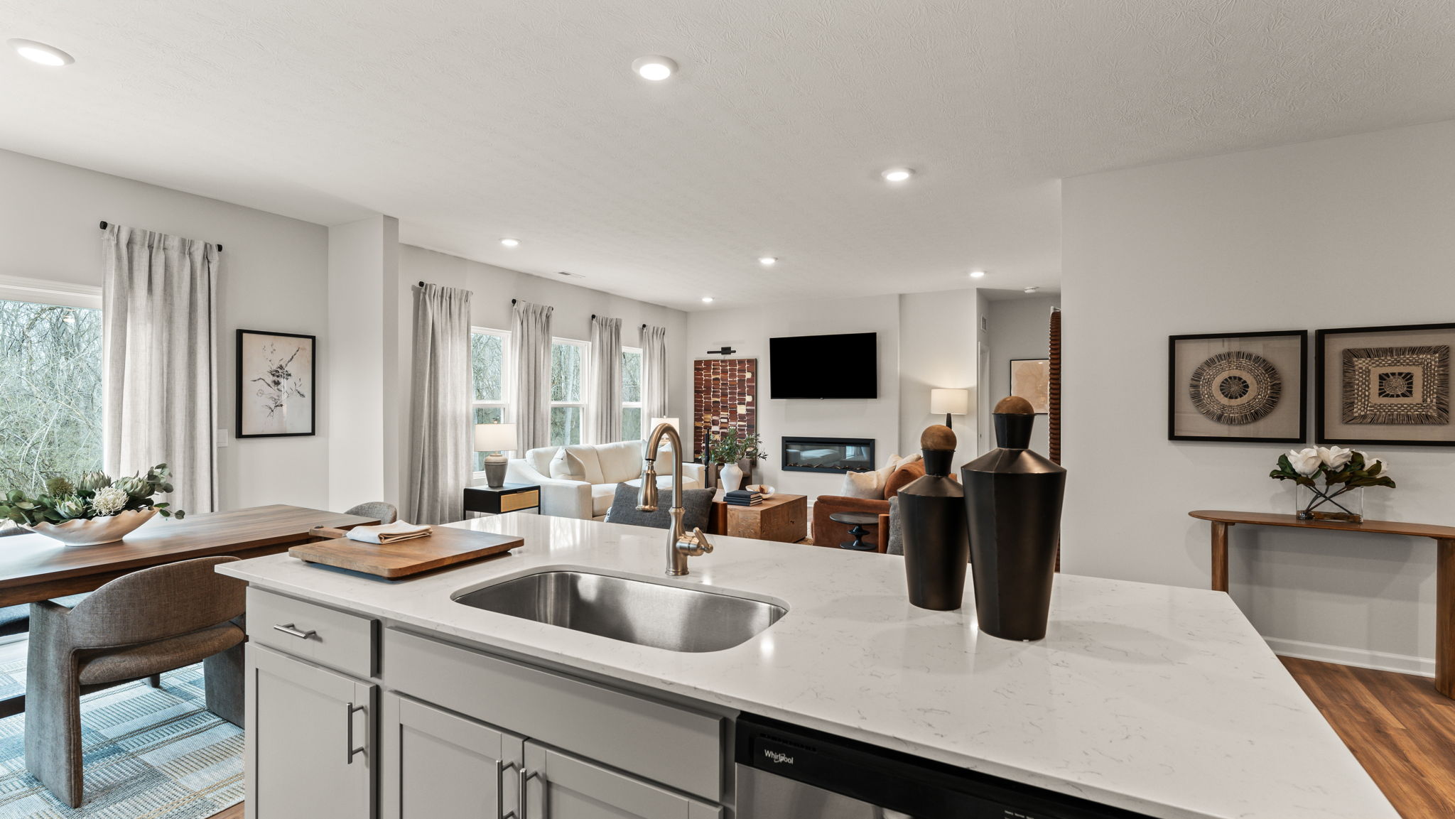 Perspective image of a warm, cozy kitchen and living room with brown wood tones, electric fireplace, and chairs