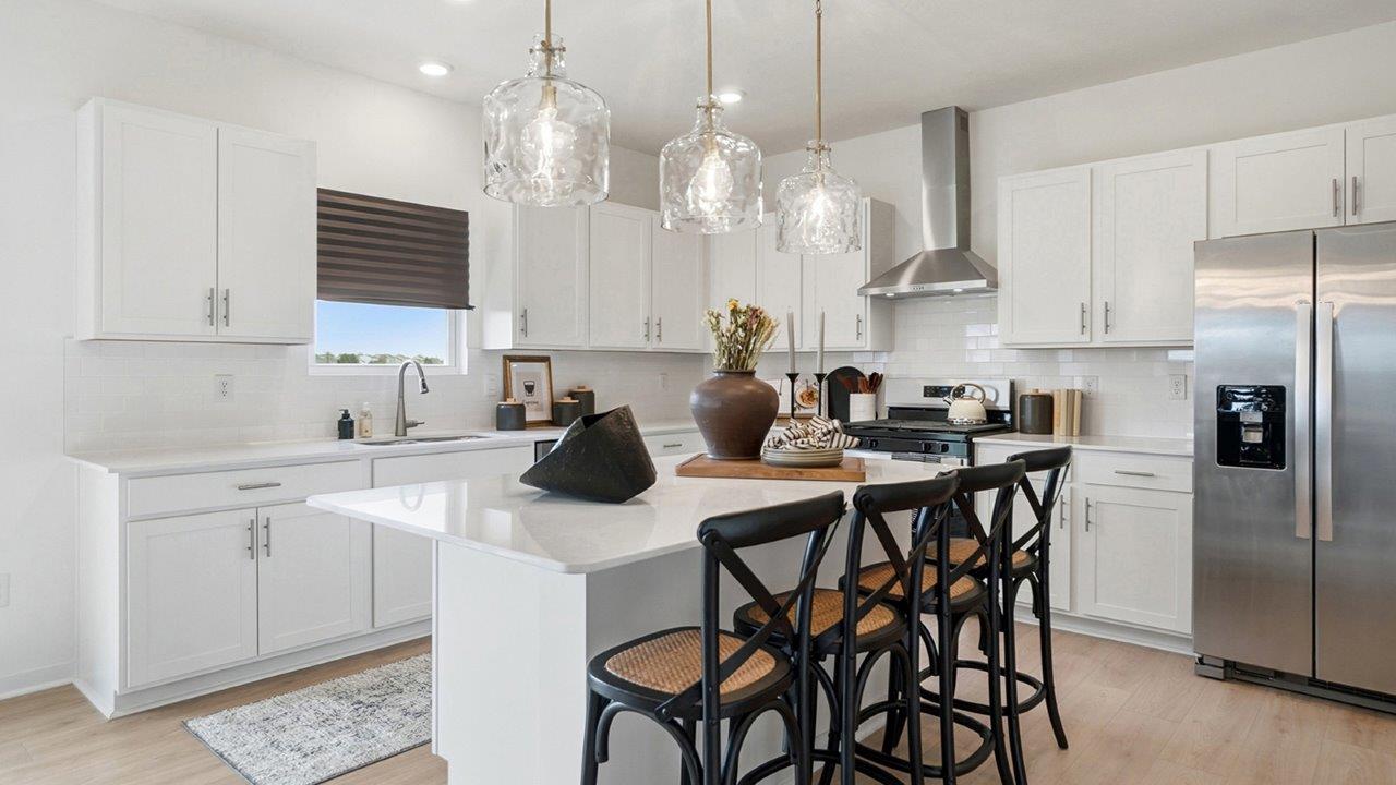 Bright white kitchen with a large island, four black counter stools, three glass pendant lights, stainless steel appliances, and a window above the sink.