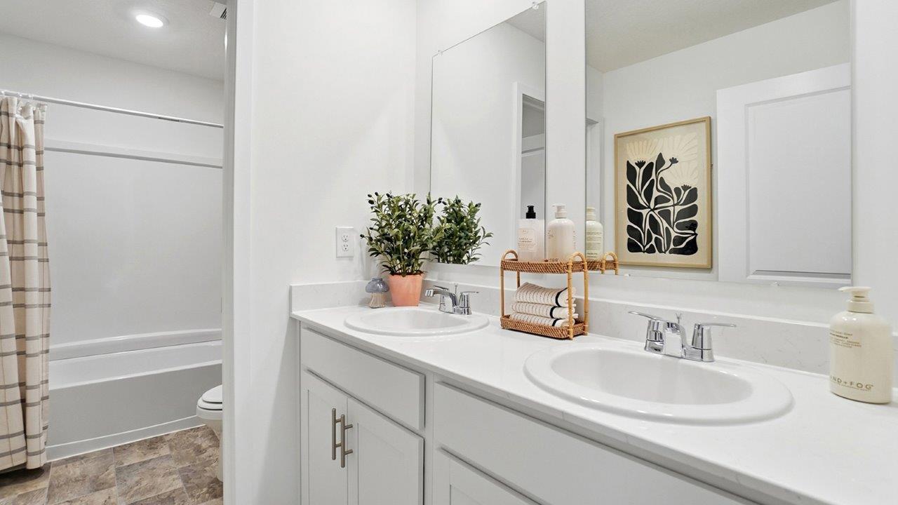 White double-sink bathroom vanity with large mirrors, chrome fixtures, greenery, and a tub visible at left.