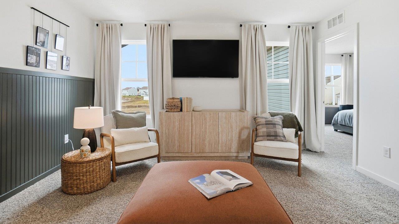 Loft seating area with two chairs facing a large orange ottoman and wall-mounted television between tall windows.