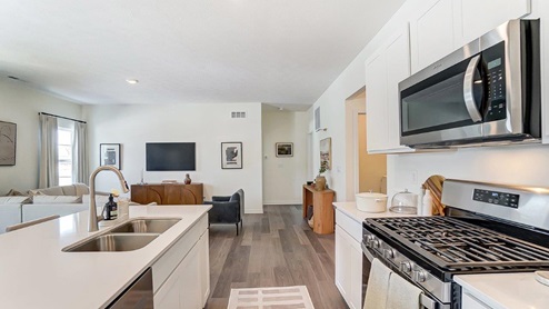 kitchen island with built in sink, white cabinetry with stainless microwave and range, great room