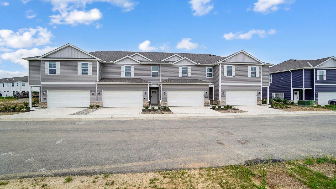 A two-story home with tan siding, a two-car garage, two windows with shutters, and a white front door with a small transom.