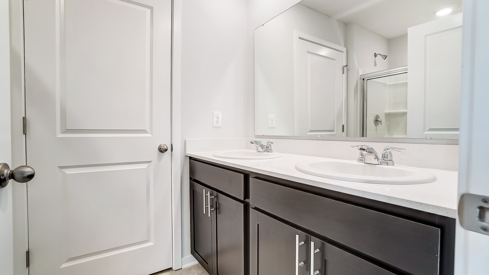 Ensuite restroom with a double vanity sink, white countertops, ebony shaker-style cabinets, and a white walk-in shower with a sliding glass door.