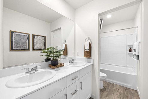 Amber Lynn Estates Plan 1811 Twin sinks with white countertops, white cabinets, wide mirror, hand towel ring, towel bar, toilet, and shower.