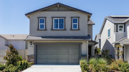 Two story home with tan siding and stone detailing, covered patio, and two car garage