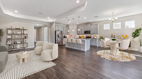 Inside of the Shelby model dining and living area. Wood tile floors with grey walls. Dining area contains a four-person dining table with hanging light fixture above it. Living area with furniture can be seen in the background as well as the hallway to the front door. Kitchen can also be seen in the background with grey cabinets and white countertops with silver hardware. Kitchen contains kitchen island with bar-style seating and stainless-steel appliances.