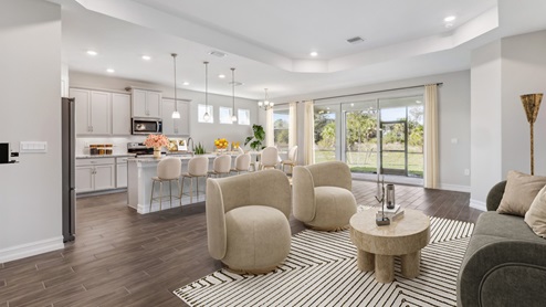 Inside of the Shelby model living area and kitchen. Kitchen can be seen in the background with grey cabinets and white countertops. Sliding glass door to the back patio can be seen in the background.