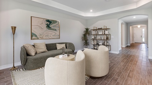 Inside of the Shelby model living room. Wood tile floors with grey walls. Room contains couch, lamp, coffee table, two armchairs, and shelf with various nick-nacks. In the background the hallway to the bedrooms and front door can be seen.