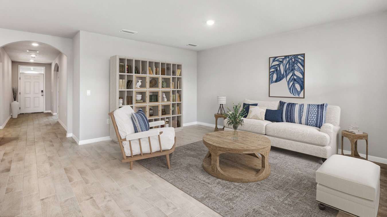 Inside of the Aria living room. Plank tile floors and off-white walls. Contains one armchair, small couch, ottoman, coffee table, bookshelf, and two small side tables on either side of couch. Living area connects to the kitchen as well as hallway to the front door.