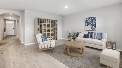 Inside of the Aria living room. Wood tile floors and off-white walls. Contains one armchair, small couch, ottoman, coffee table, bookshelf, and two small side tables on either side of couch. Living area connects to the kitchen as well as hallway to the front door.