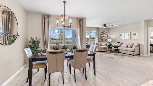 The wooden dining table is set for a family of six. There is a chandelier on top and a mirror on the wall behind. The Dining space is well lit from the patio doors. There are curtains on either side of the patio doors.