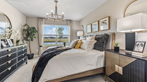 The Master Bedroom is carpeted with grey painted walls. There is a window with a view of the back of the home. There are side tables on either side of the freshly made bed. There is a chandelier above the bed as well. Here you can see the dresser for clothes across from the bed and the plant in the corner.