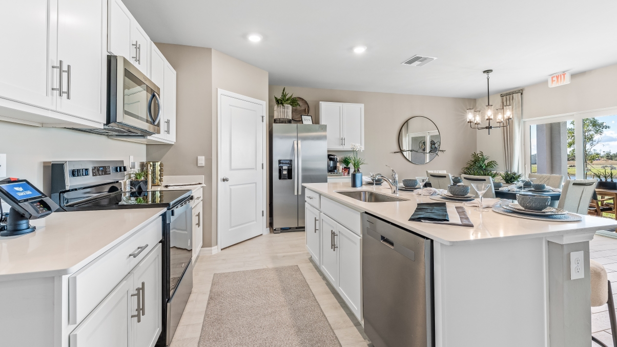 Inside the Cali Model kitchen. This home includes tiles flooring, stainless steel appliances and white cabinetry with silver fixtures. The bar is great for hosting people in the kitchen. You can see the dining room and patio from here. Theres a clear view outside.