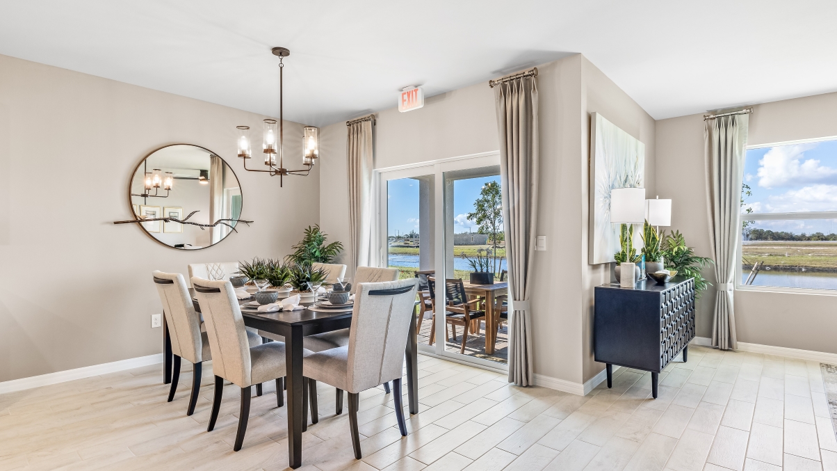 The wooden dining table is set for a family of six. There is a chandelier on top and a mirror on the wall behind. The Dining space is well lit from the patio doors. There are curtains on either side of the patio doors.