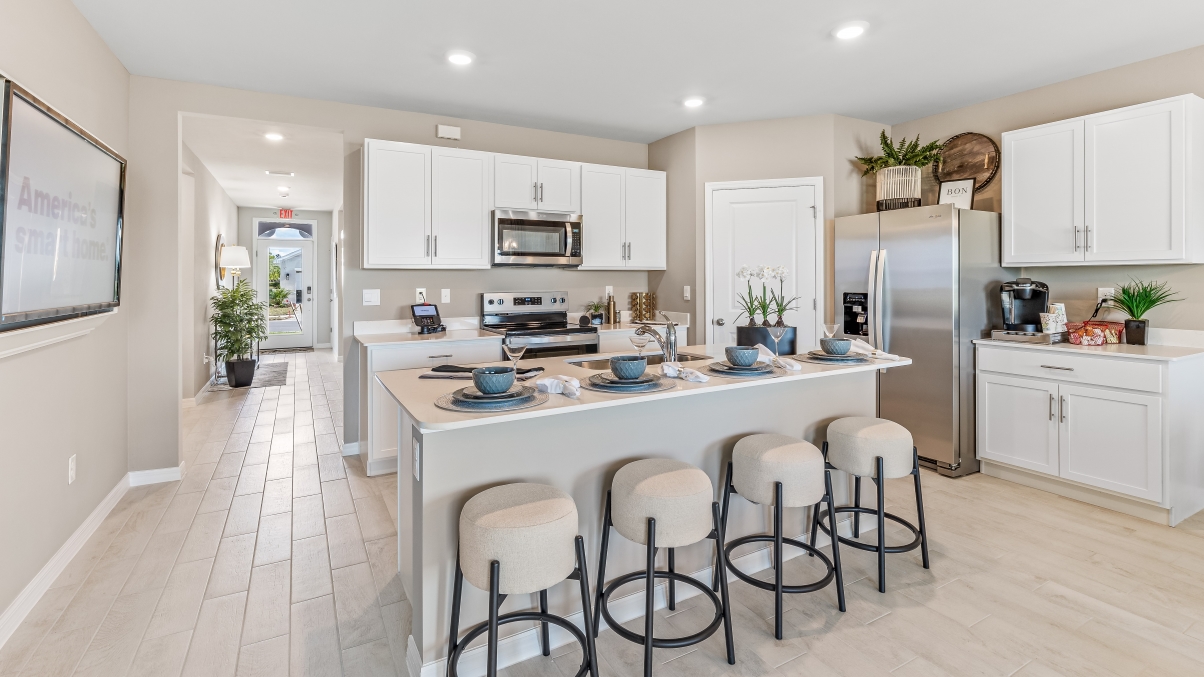 Inside the Cali Model kitchen. You can see the entrance hallway from the door. This home includes tiles flooring, stainless steel appliances and white cabinetry with silver fixtures. The bar is great for hosting people in the kitchen.