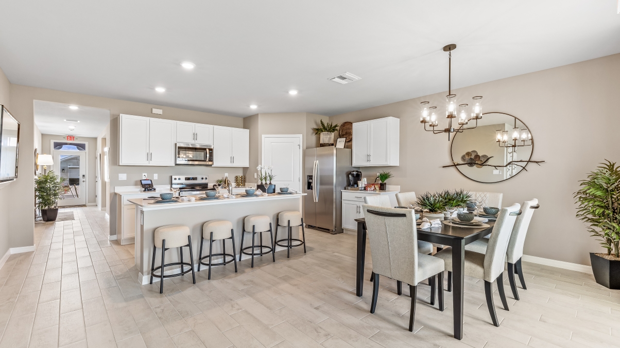 Inside the Cali Model kitchen and Dining room. You can see the entrance hallway from the door. This home includes tiles flooring, stainless steel appliances and white cabinetry with silver fixtures. The Dining space is well lit from the patio doors.