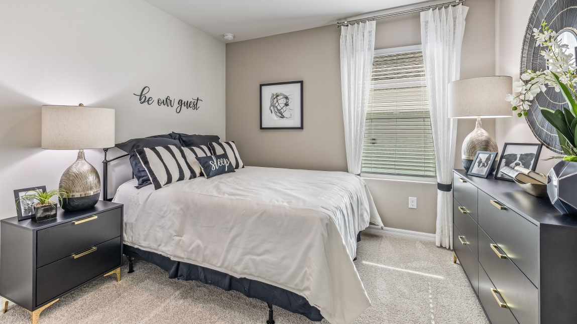 The second bedroom has a full size beds with a modern side table. There are white curtains on the window, a mirror on the wall and a dresser are across from the bed. There is small art on the wall..