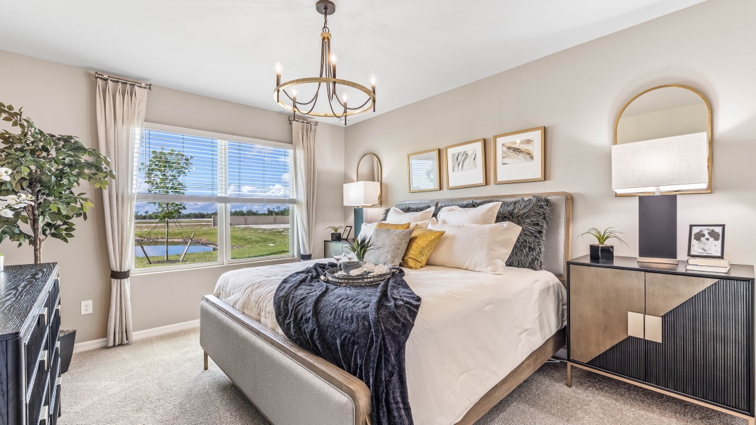 The Master Bedroom is carpeted with grey painted walls. There is a window with a view of the back of the home. There are side tables on either side of the freshly made bed. There is a chandelier above the bed as well. Here you can see the dresser for clothes across from the bed and the plant in the corner.
