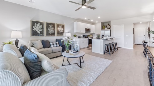 Inside the Bayside model living room. You can see the entrance of the townhome as well as the kitchen from here. There is enough room for a medium sized couch and 2 chairs, a coffee table, there is a rug on the floor and artwork on the walls. Theres a celing fan as well.