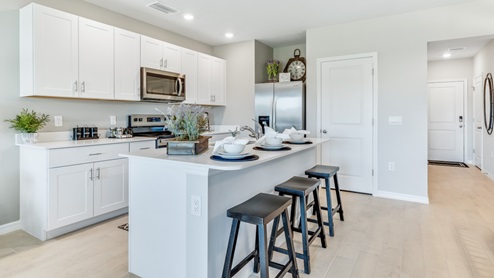 The kitchen has modern solid white cabinetry and stainless steel appliances. There is a pantry and bar seating. You can see the front door from this angle.