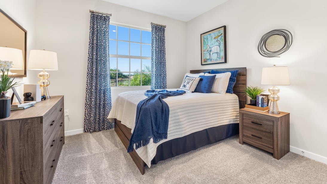Inside the Lantana model bedroom 1. Contains a bed with a side table, and a dresser across from the bed. Also contains a window.