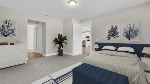 Inside of the Delray B model bedroom one. Grey carpet with off-white walls. Room contains one bed with small nightstands on either side, white dresser, and a bench at the end of the bed. In the background there is a door leading to the kitchen area as well as another hallway leading to a closet and other room.