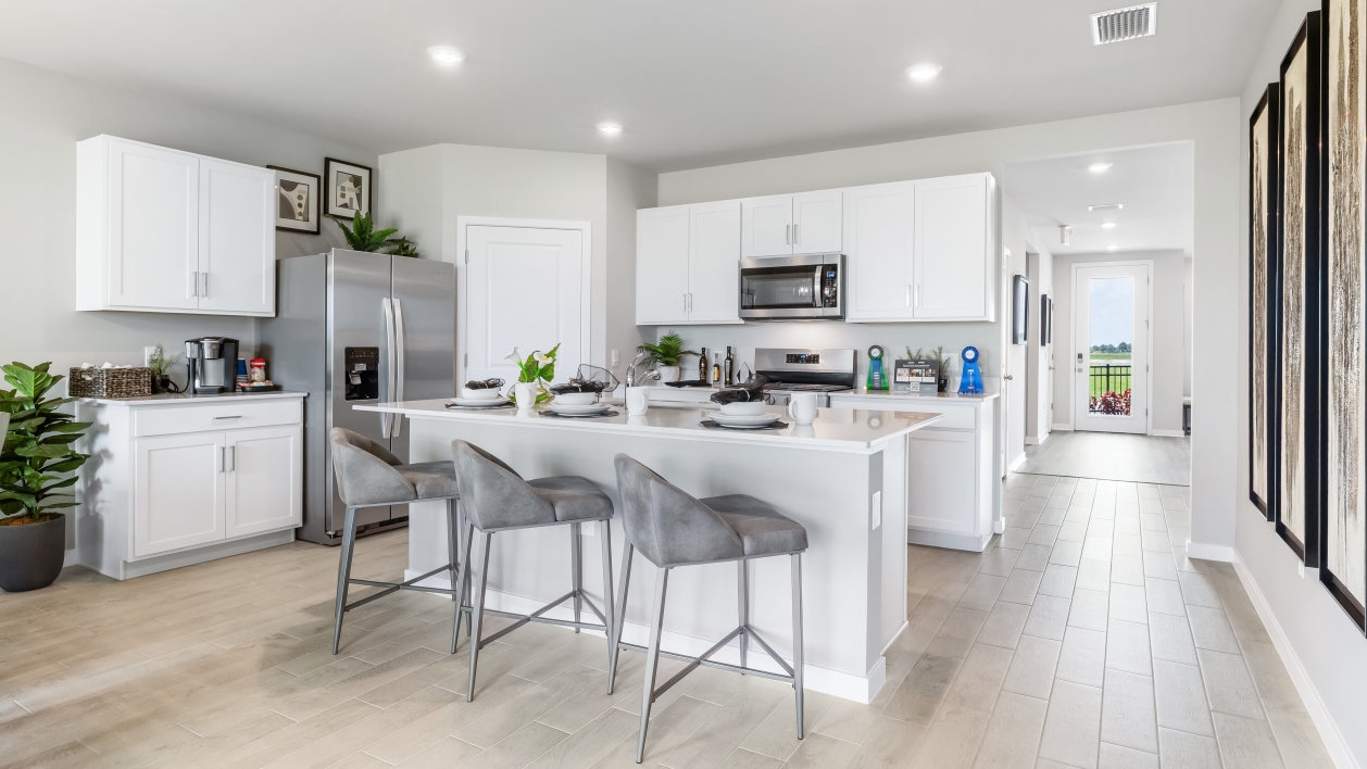 Inside of the Cali model kitchen. Grey Plank tile flooring and grey walls. Kitchen has white cabinets with silver hardware, white countertops, and stainless-steel appliances. There is also a kitchen island with bar-style seating. In the background there is the hallway that connects to the front door and other rooms.