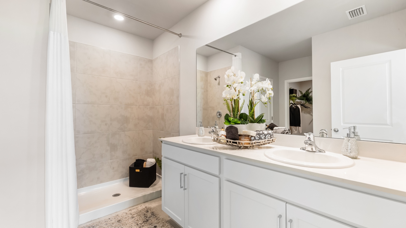 Inside of the Cali model bathroom. Shower has cream tiles. Dual vanity with cream countertops and cabinets with silver hardware.