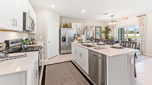 Inside the Cali Model kitchen. This home includes tiles flooring, stainless steel appliances and white cabinetry with silver fixtures. The bar is great for hosting people in the kitchen. You can see the dining room and patio from here. Theres a clear view outside.