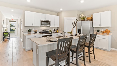 Inside the Cali Model kitchen. You can see the entrance hallway from the door. This home includes tiles flooring, stainless steel appliances and white cabinetry with silver fixtures. The bar is great for hosting people in the kitchen.