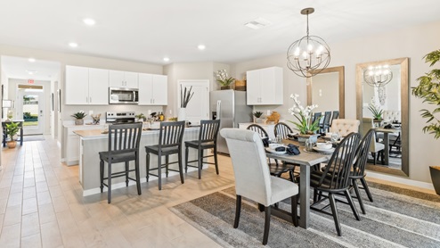 Inside the Cali Model kitchen and Dining room. You can see the entrance hallway from the door. This home includes tiles flooring, stainless steel appliances and white cabinetry with silver fixtures. The Dining space is well lit from the patio doors.