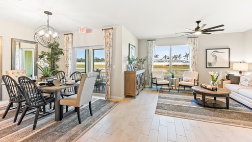 The wooden dining table is set for a family of six. There is a chandelier on top and a mirror on the wall behind. The Dining space is well lit from the patio doors. There are curtains on either side of the patio doors. You can also see the living room from here