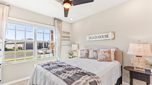 The Master Bedroom is carpeted with grey painted walls. There is a window with a view of the back of the home. There are side tables on either side of the freshly made bed. There is a chandelier above the bed as well. Here you can see the dresser for clothes across from the bed and the plant in the corner.