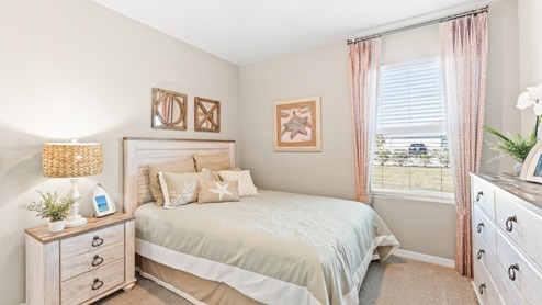 The second bedroom has a full size beds with a modern side table. There are white curtains on the window, a mirror on the wall and a dresser are across from the bed. There is small art on the wall..